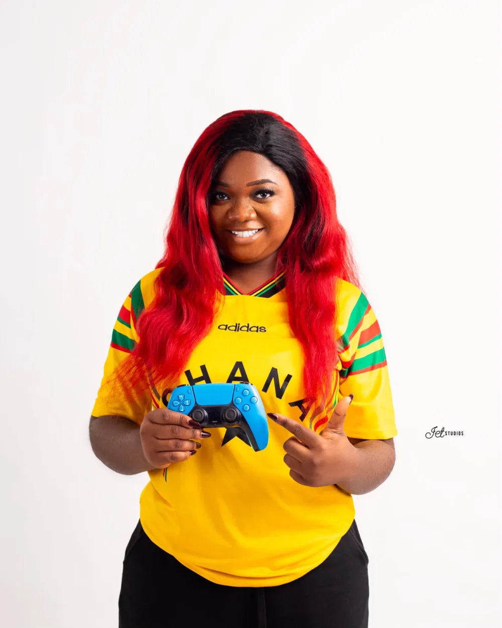 Vanessa Selase Azoumaro in a Ghana national team jersey, pointing at the blue PlayStation controller in her hand.